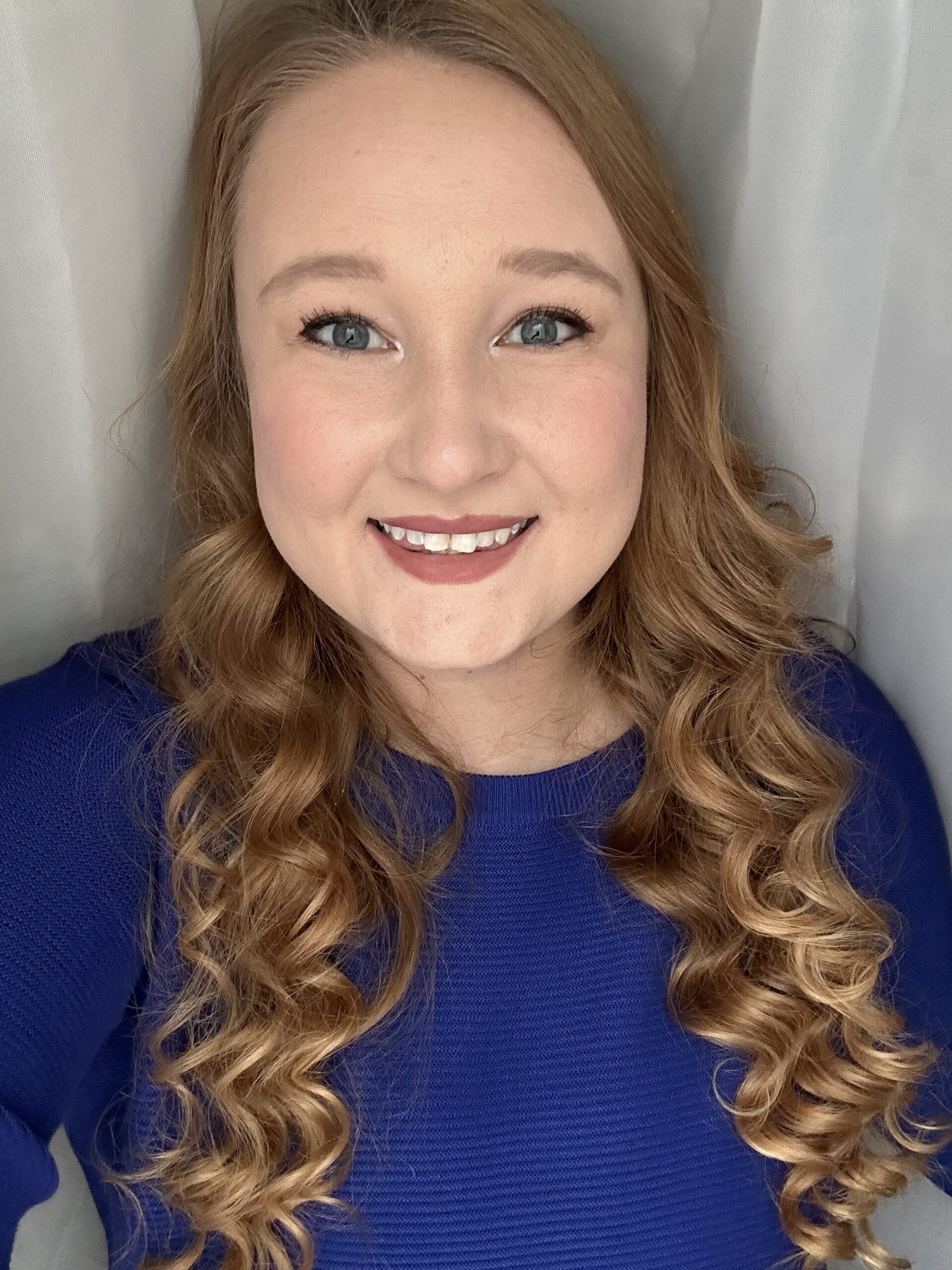 Jessika stands in front of a sheer white curtain smiling, with curly hair and wearing a blue sweater.