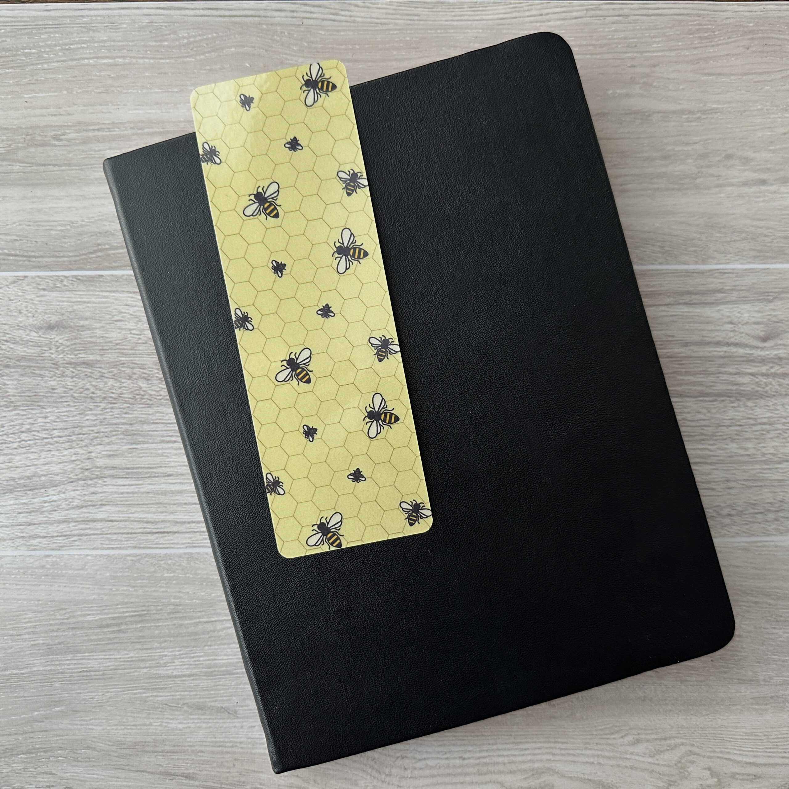 A bookmark sporting a pattern of bees with a yellow background rests upon a black notebook on a table.