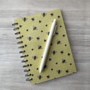 A wire-o bound notebook with a bee pattern and yellow background. A white pen rests across the cover.