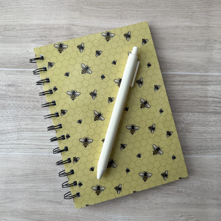 A wire-o bound notebook with a bee pattern and yellow background. A white pen rests across the cover.