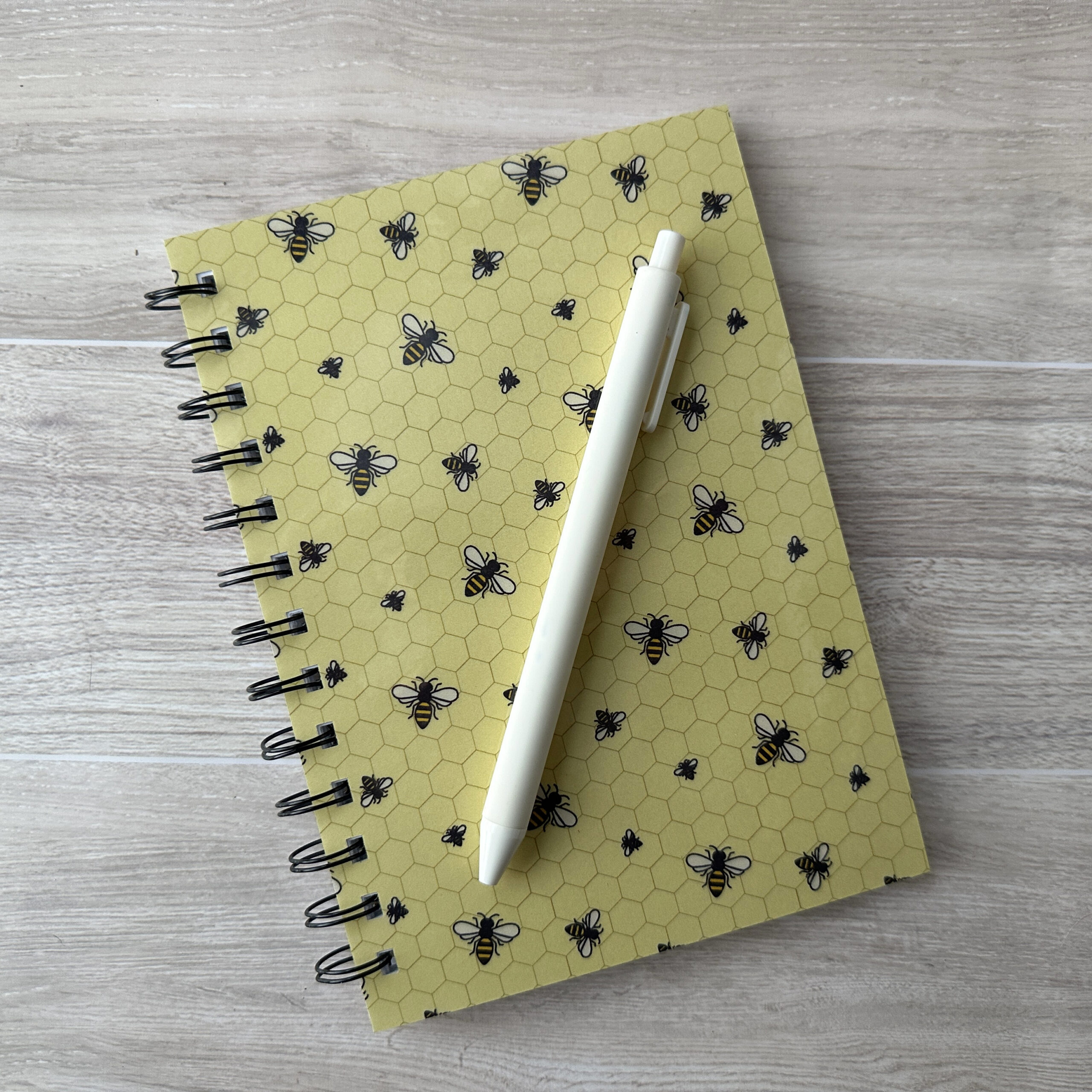A wire-o bound notebook with a bee pattern and yellow background. A white pen rests across the cover.