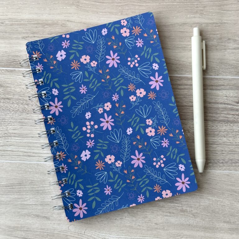 A wire-o notebook with a blue floral pattern sits on a table with a white pen