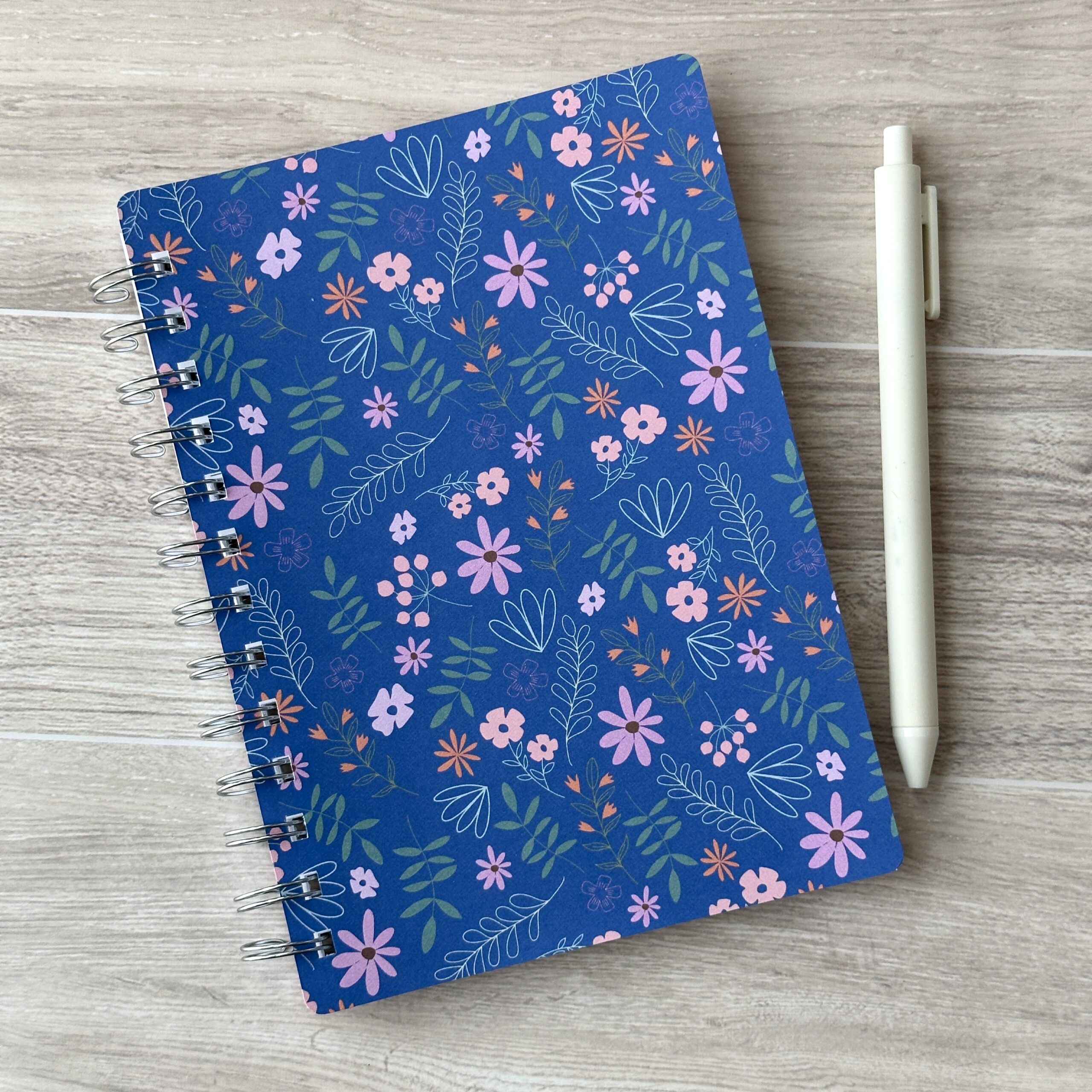 A wire-o notebook with a blue floral pattern sits on a table with a white pen