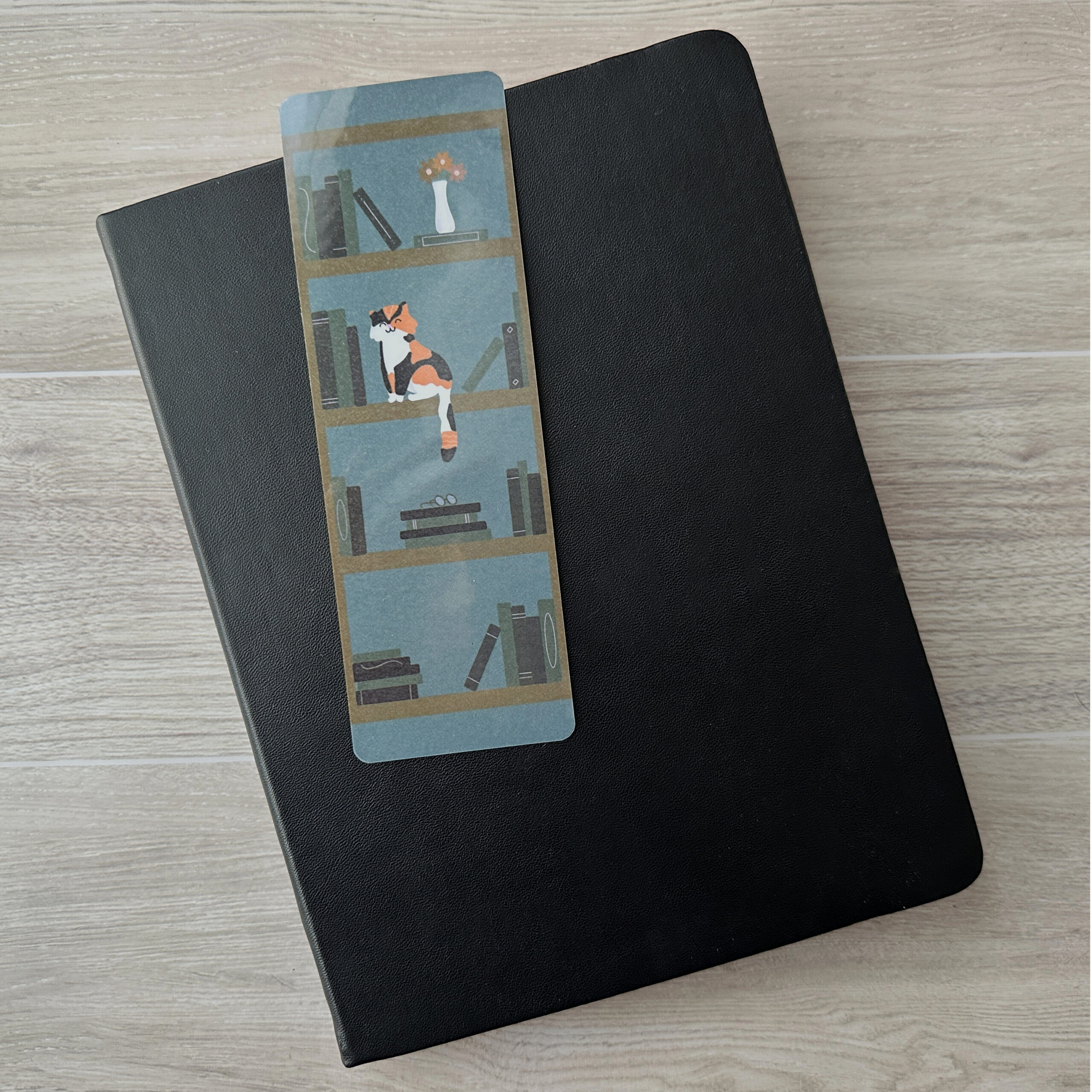 A bookmark sporting a bookshelf and a cat with a blue background rests upon a black notebook on a table.