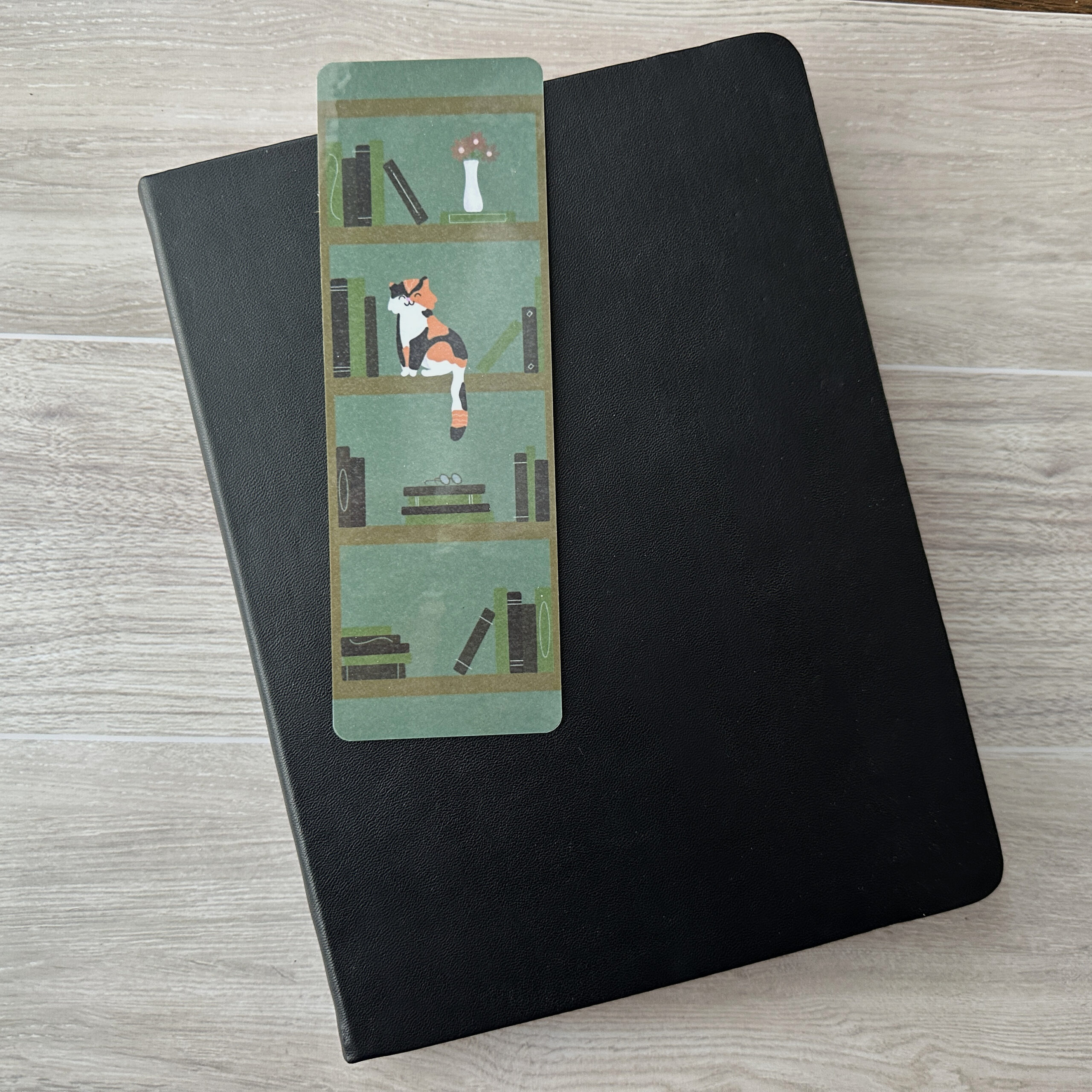 A bookmark sporting a bookshelf and a cat with a green background rests upon a black notebook on a table.