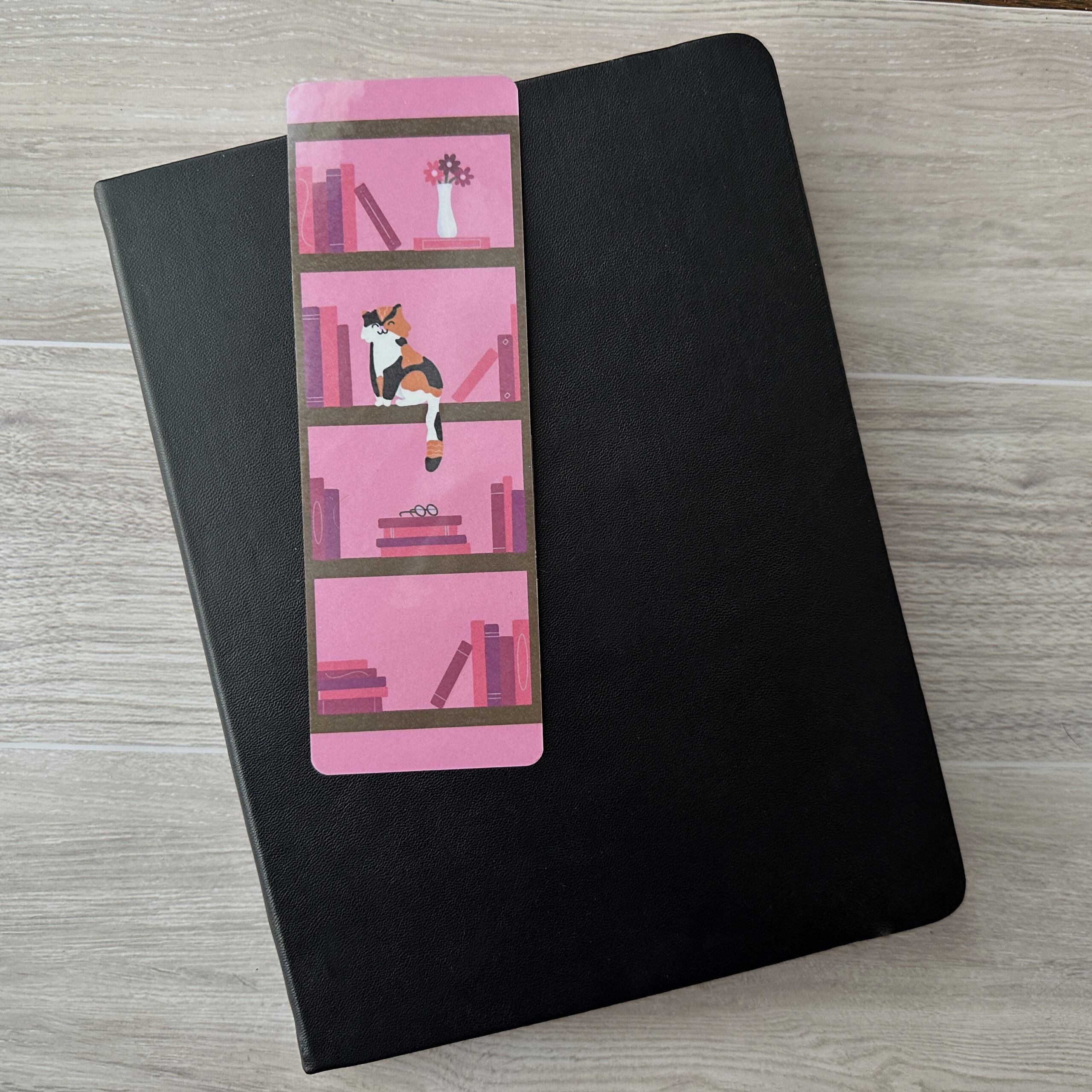 A bookmark sporting a bookshelf and a cat with a Pink background rests upon a black notebook on a table.