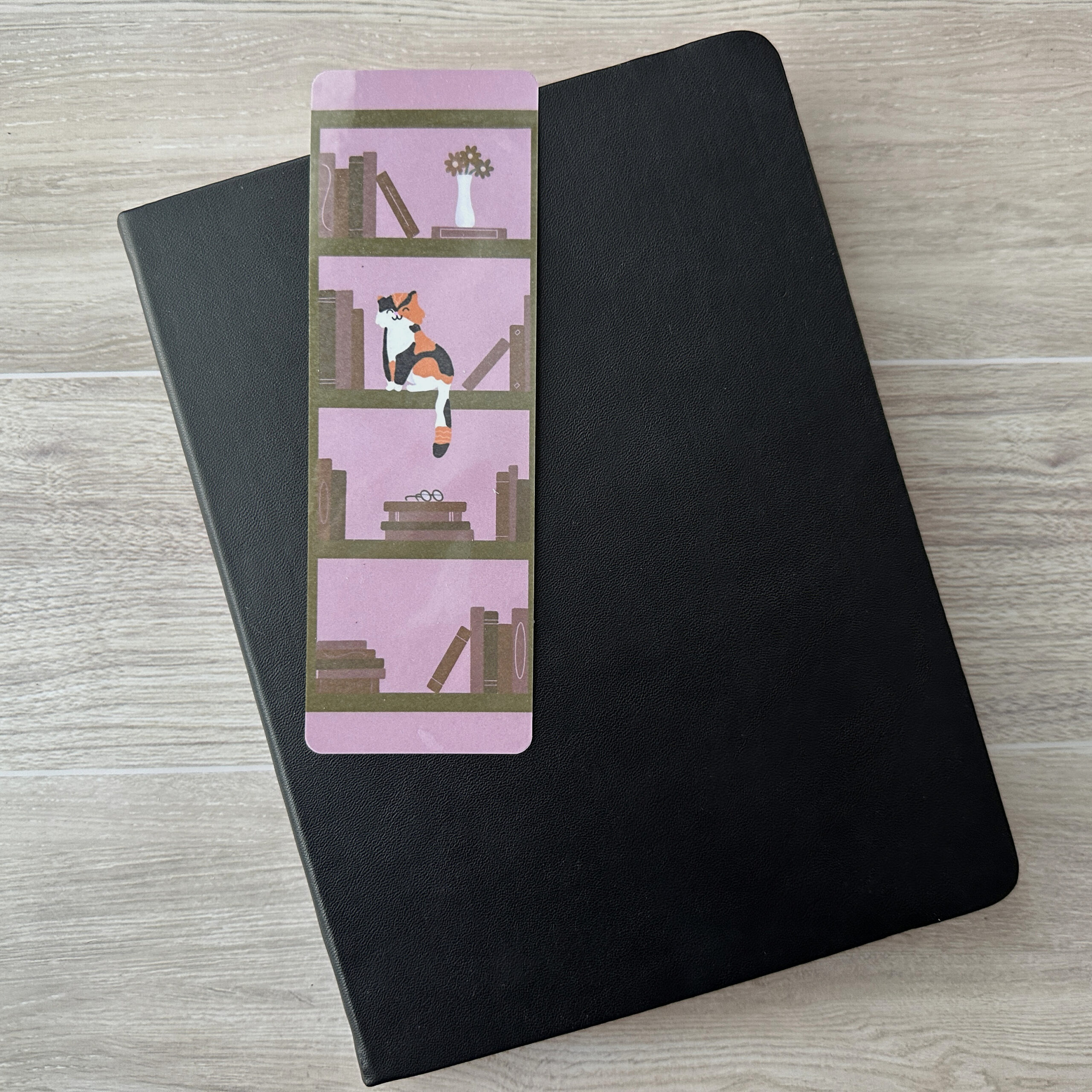 A bookmark sporting a bookshelf and a cat with a purple background rests upon a black notebook on a table.