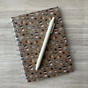 A wire-o bound notebook with assorted coffee items in a pattern on the cover with a medium brown background. A white pen rests across the cover