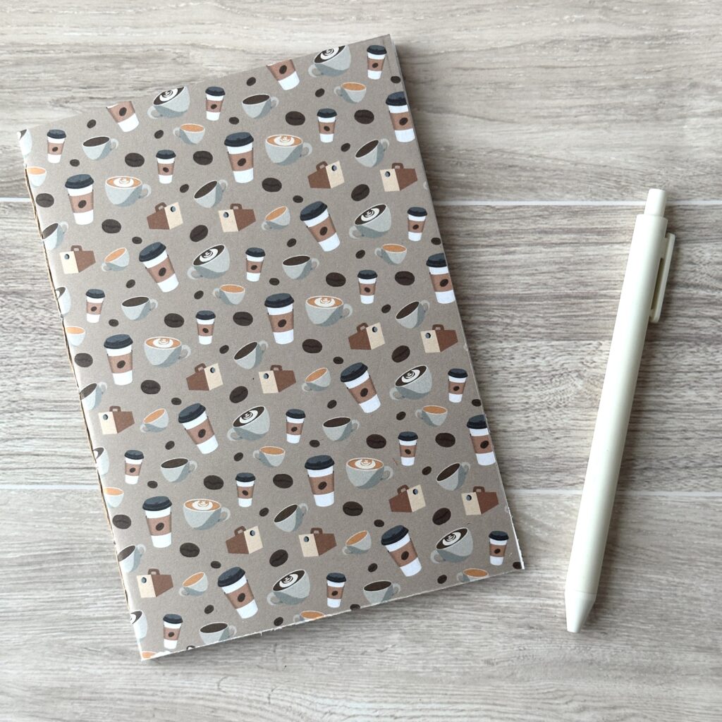 A notebooklet with an assorted coffee items pattern sits on a table with a white pen