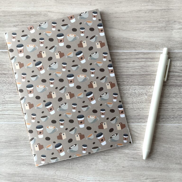 A notebooklet with an assorted coffee items pattern sits on a table with a white pen
