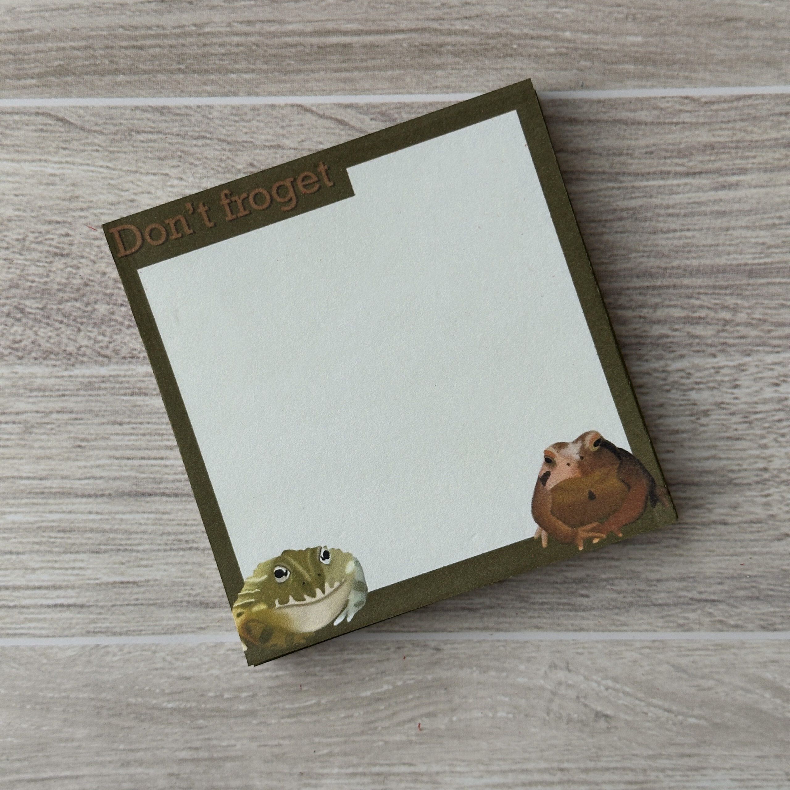 A square sticky notepad with a frog in each bottom corner and the statement"dont froget" on the top left. There is a greenish-brown border.
