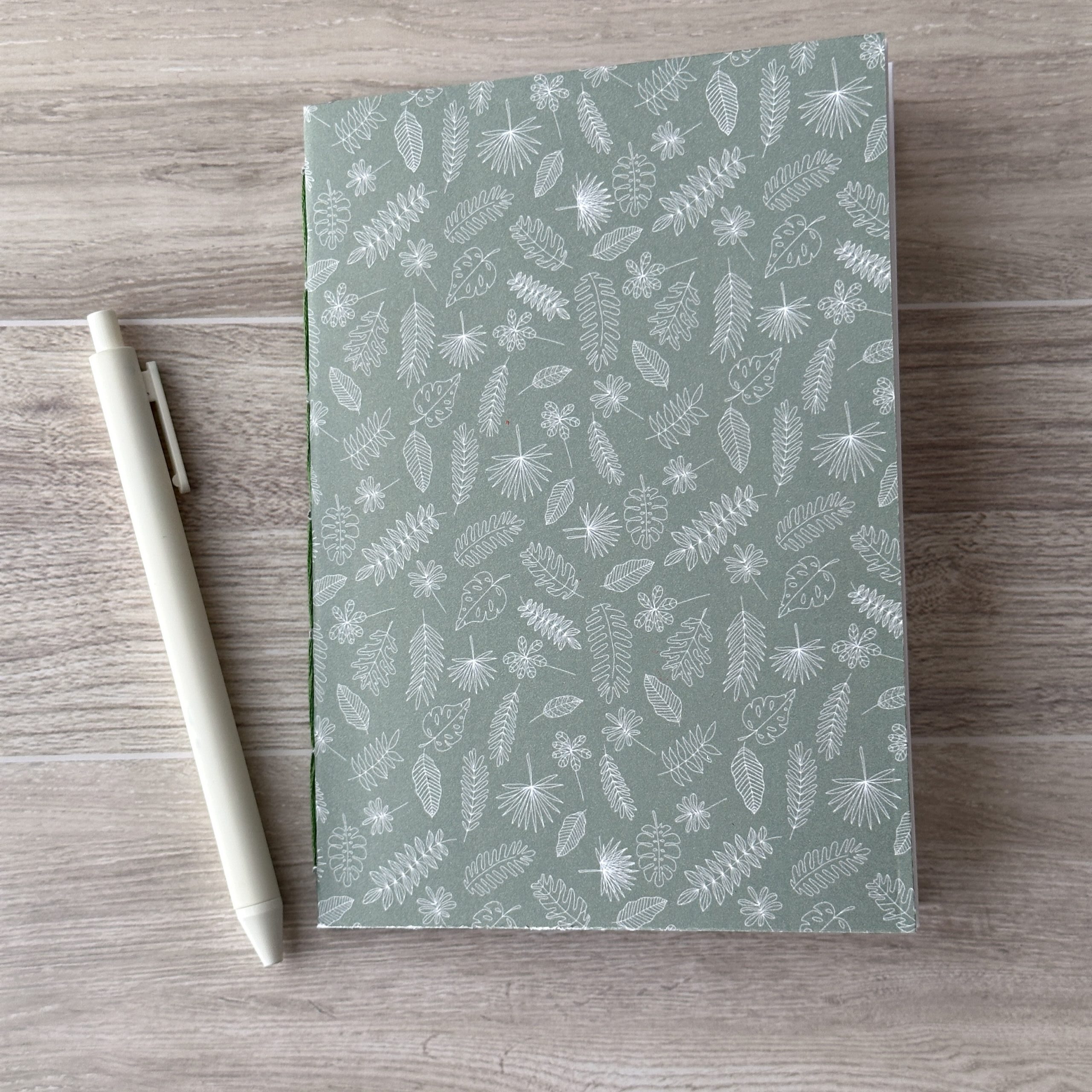 A notebooklet with an assorted leaves pattern sit on a table with a white pen