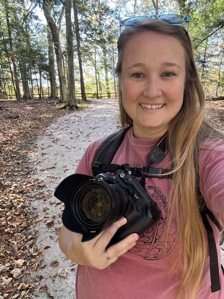 Jessika stands in the middle of a gravel path in the woods holding her camera and smiling. There are fall leaves on the ground