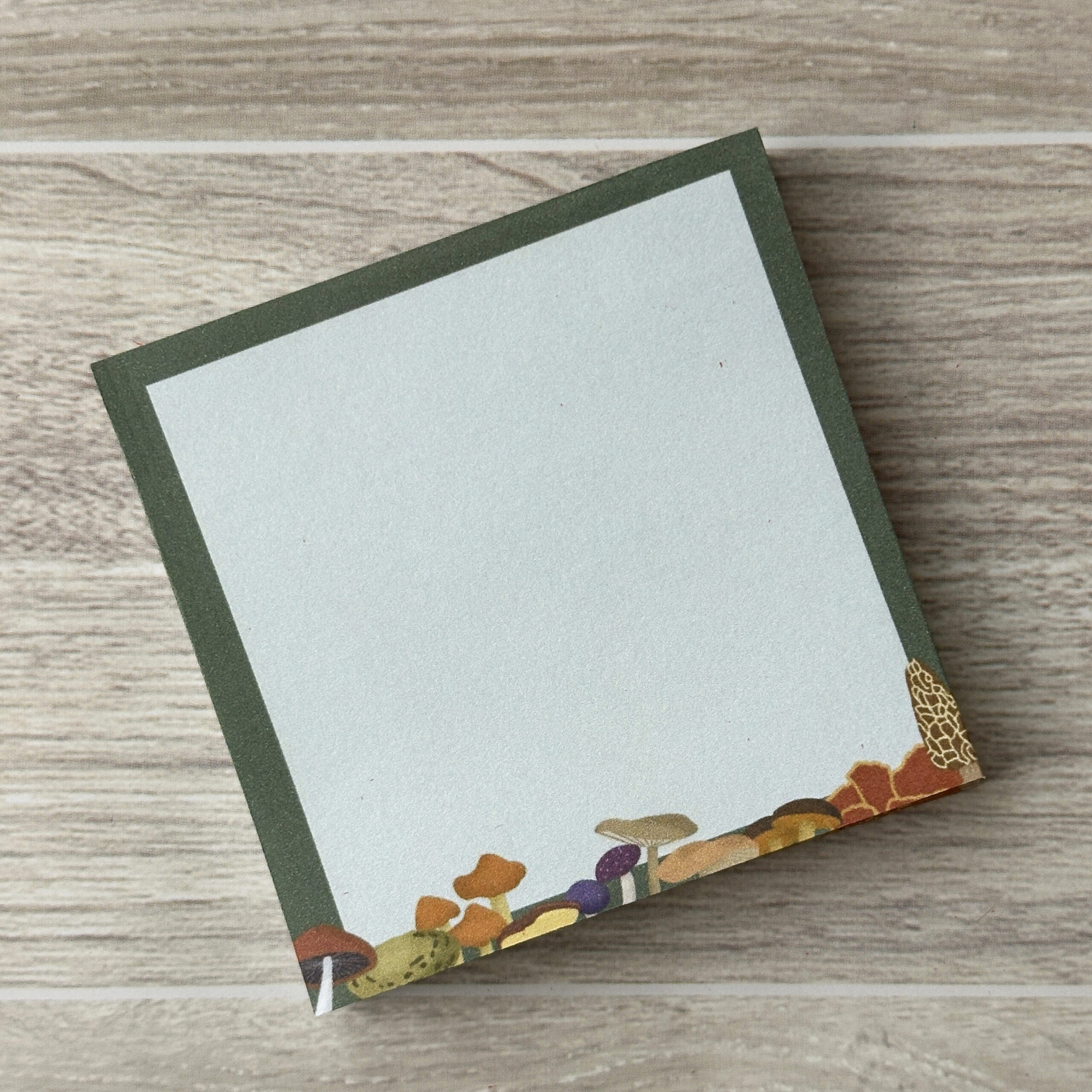 A square sticky note pad with an assortment of mushrooms along the bottom and a dark green border.
