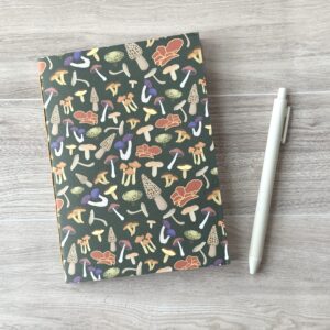 A notebooklet with an assorted mushroom pattern and green background sits on a table with a white pen