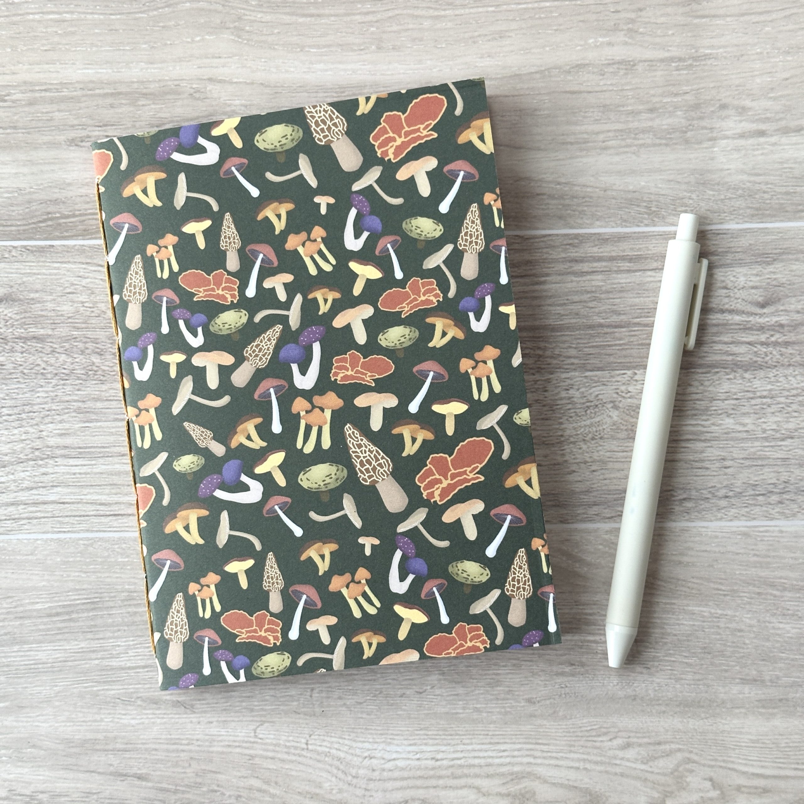 A notebooklet with an assorted mushroom pattern and green background sits on a table with a white pen