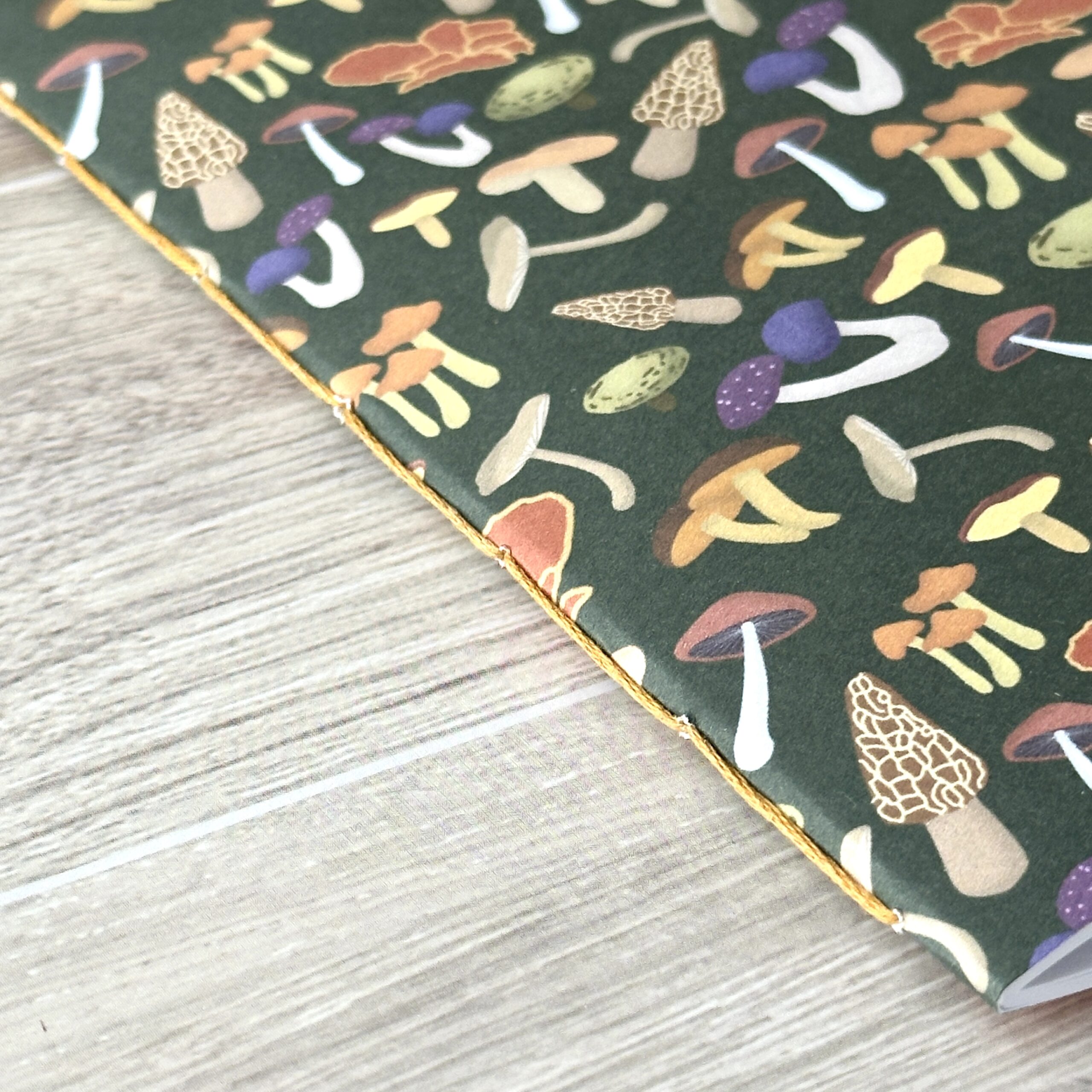 A notebooklet with an assorted mushroom pattern and green background sits on a table with a close up of the binding
