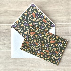 A card with an assorted mushroom pattern and dark green background with a matching envelope on a table