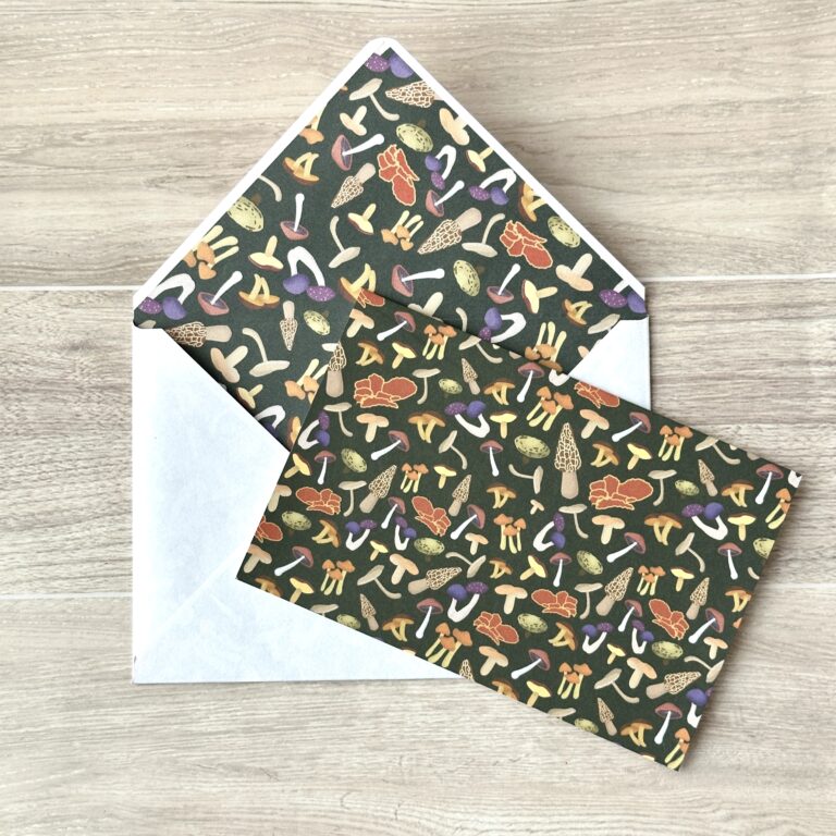 A card with an assorted mushroom pattern and dark green background with a matching envelope on a table