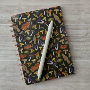 A wire-o bound notebook with an assorted mushroom patterned cover and a dark green background. A white pen sits across the cover.