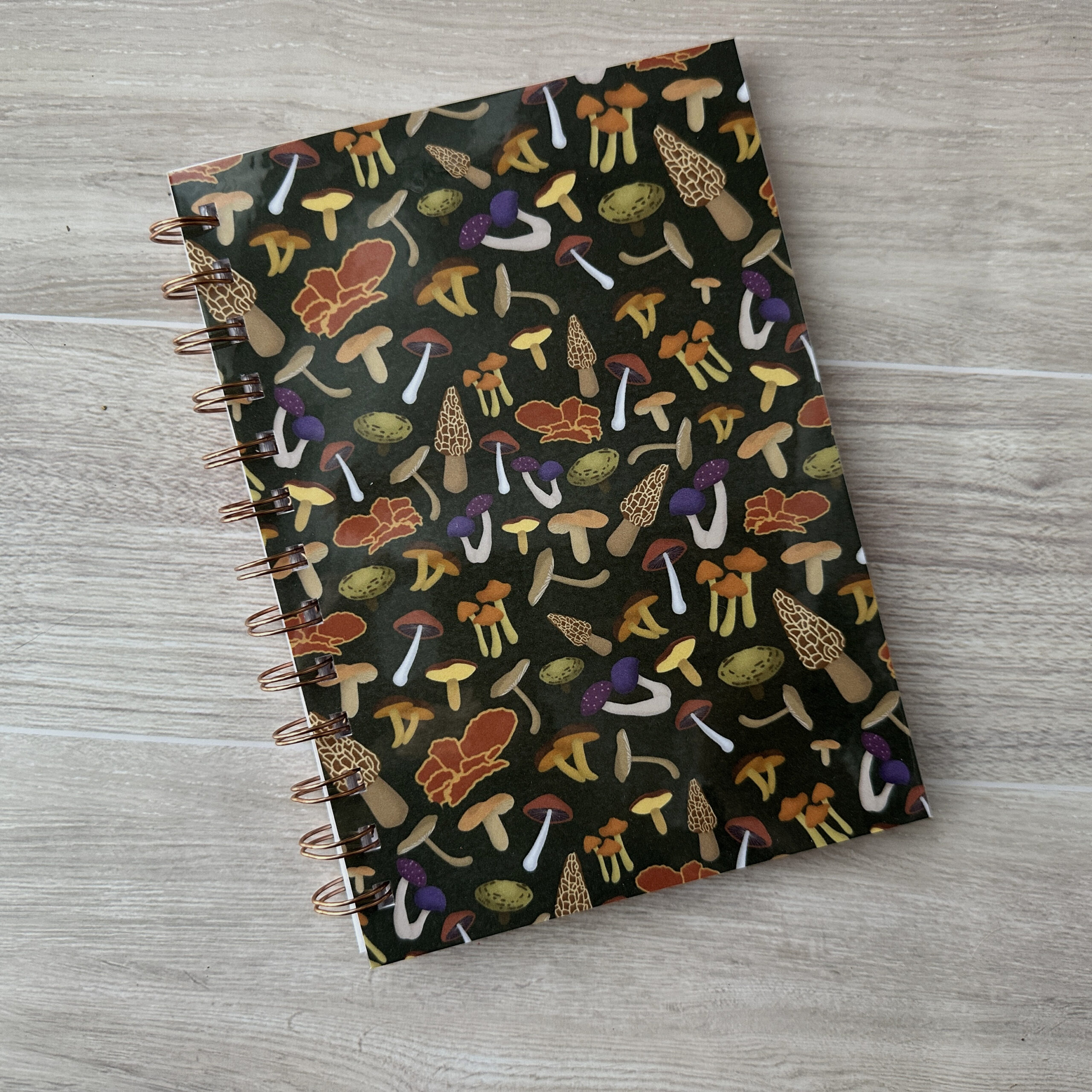 A wire-o bound notebook with an assorted mushroom patterned cover and a dark green background.