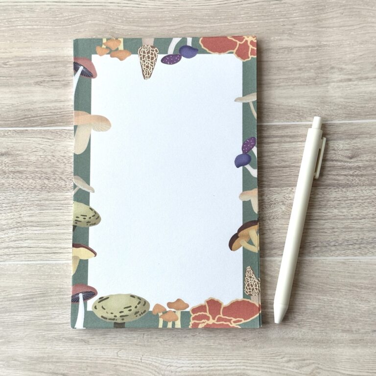 A paper pad with an assorted mushroom pattern and green background sits on a table with a white pen