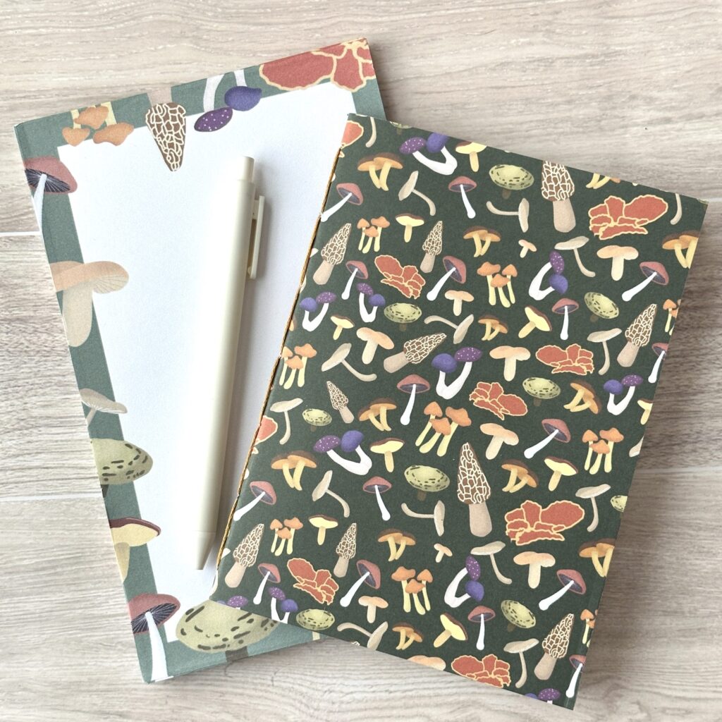 A wire-o notebook and a paper pad with an assorted mushroom pattern and green background sit on a table with a white pen