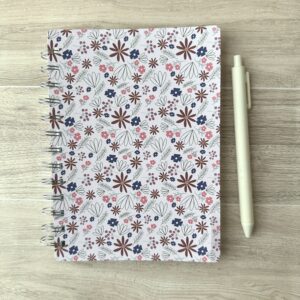 A wire-o notebook with a pink floral pattern sits on a table with a white pen