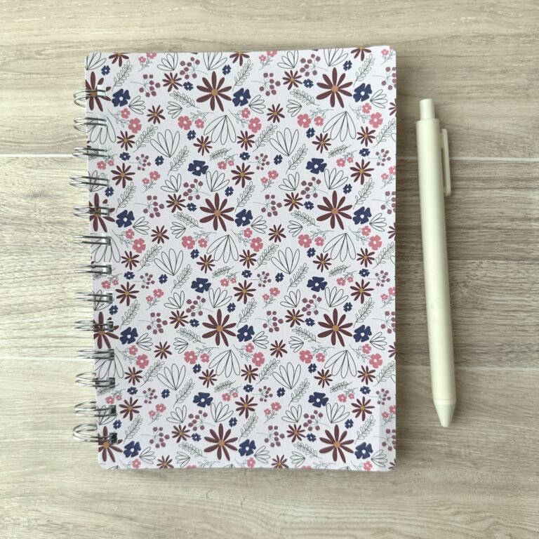 A wire-o notebook with a pink floral pattern sits on a table with a white pen
