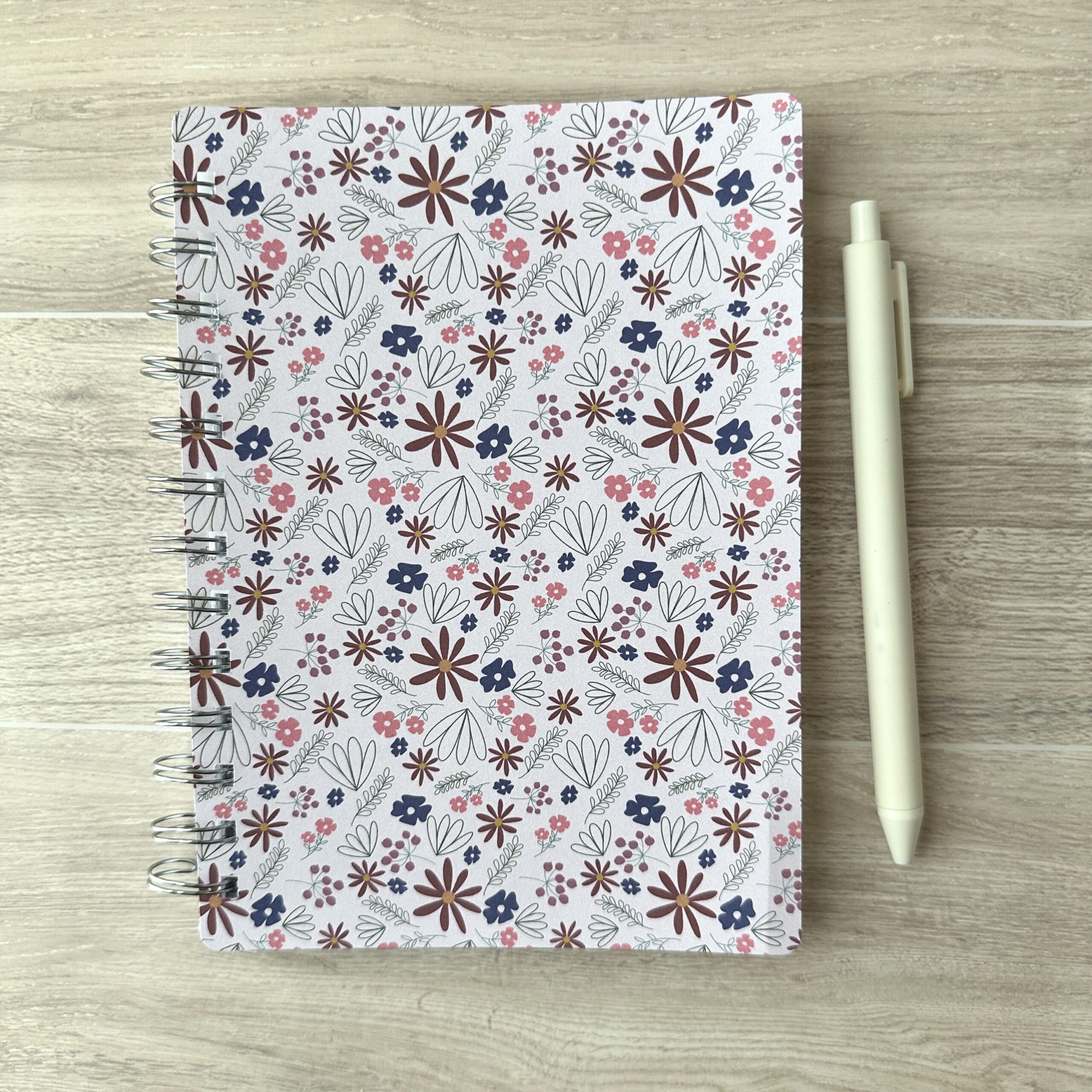 A wire-o notebook with a pink floral pattern sits on a table with a white pen