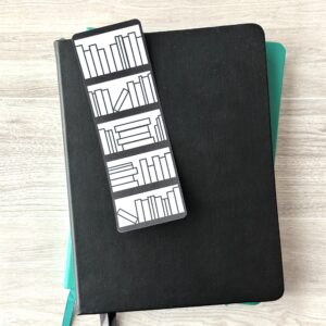 A bookmark sporting a Bookshelf with empty books on a white background rests upon a black notebook and a blue notebook on a table.