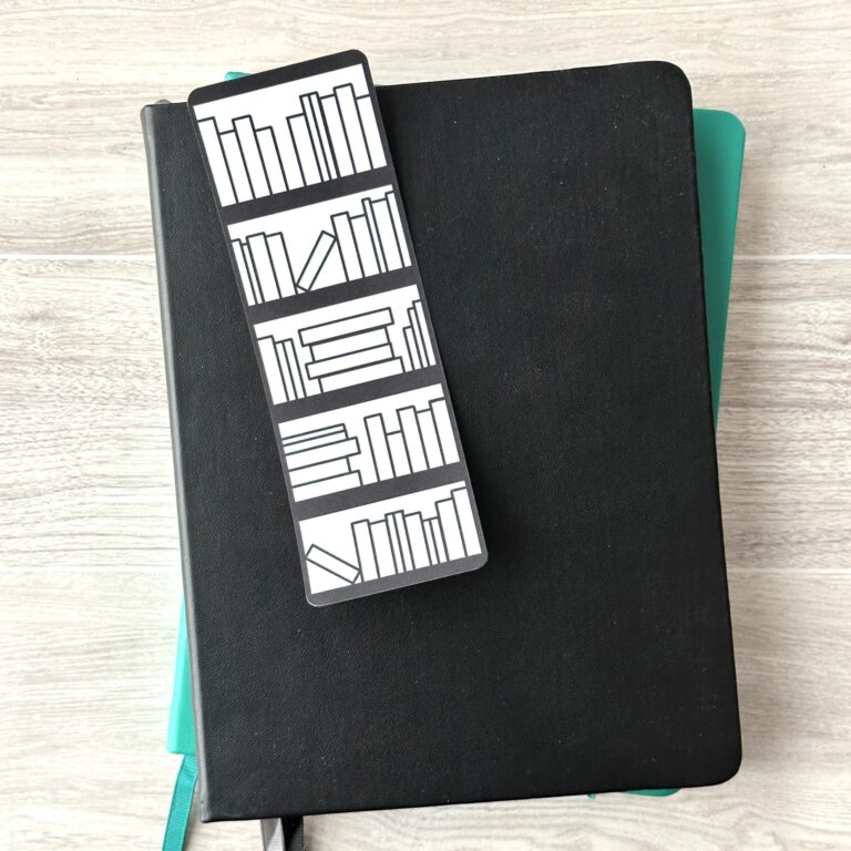 A bookmark sporting a Bookshelf with empty books on a white background rests upon a black notebook and a blue notebook on a table.