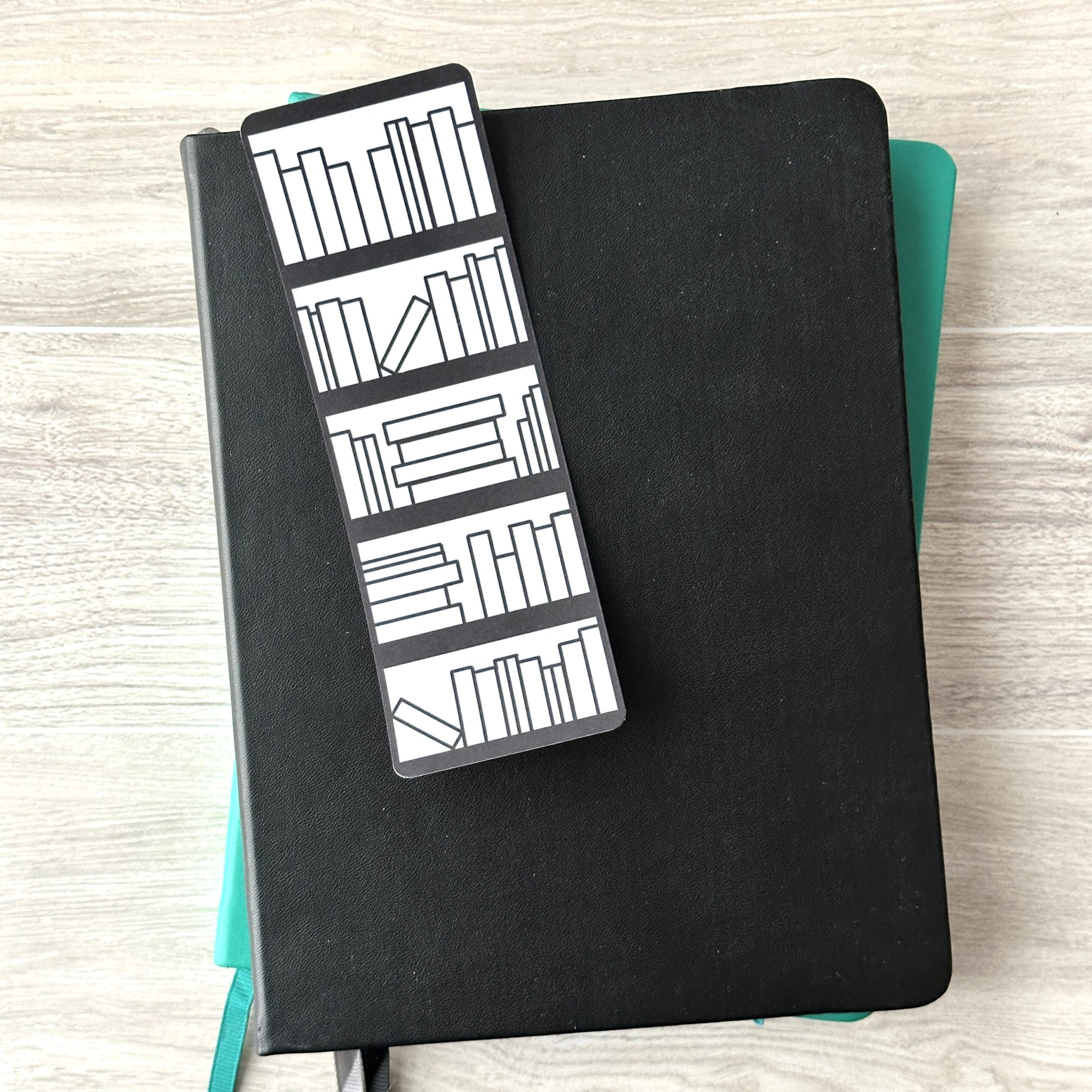 A bookmark sporting a Bookshelf with empty books on a white background rests upon a black notebook and a blue notebook on a table.