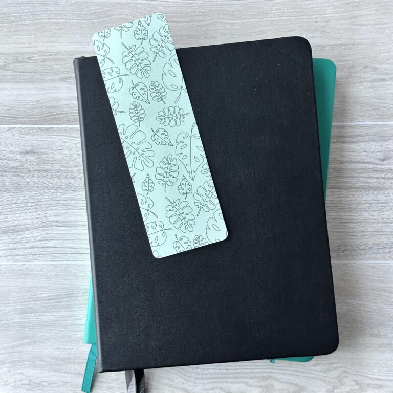 A bookmark sporting a pattern of assorted tropical leaves with a blue background rests upon a black notebook and a blue notebook on a table.