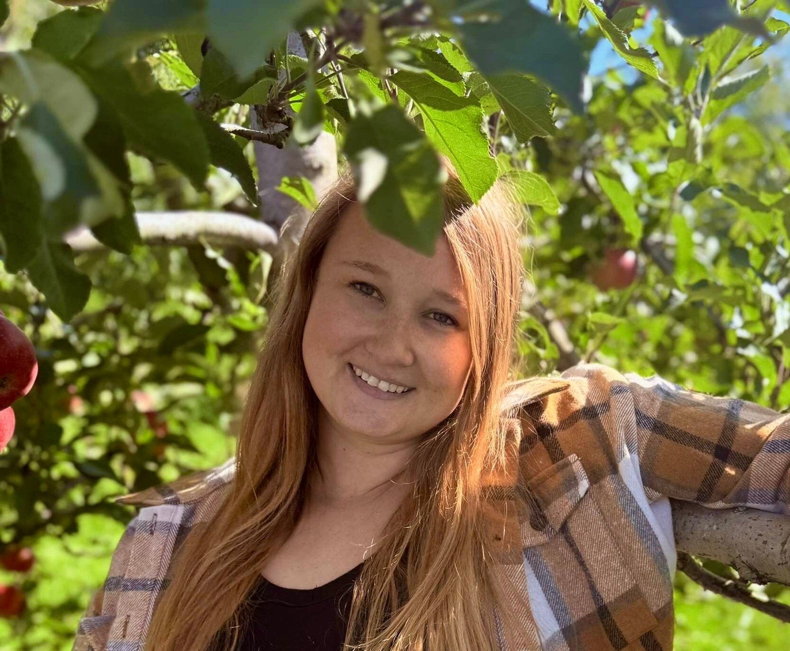Jessika stands in front of an apple tree wearing a plaid jacket
