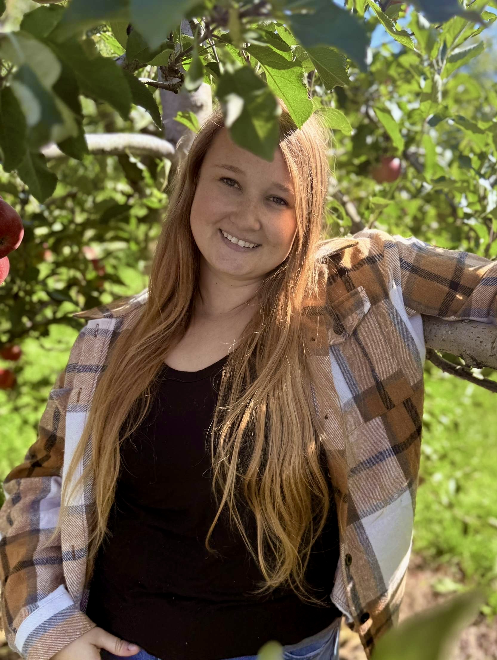 Jessika stands in front of an apple tree wearing a plaid jacket