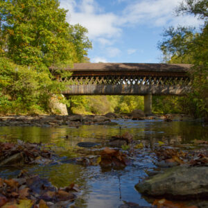 State road bridge in ashtabula viewed at the side from the river. There are fall leaves, a blue sky, and rocks in the river.
