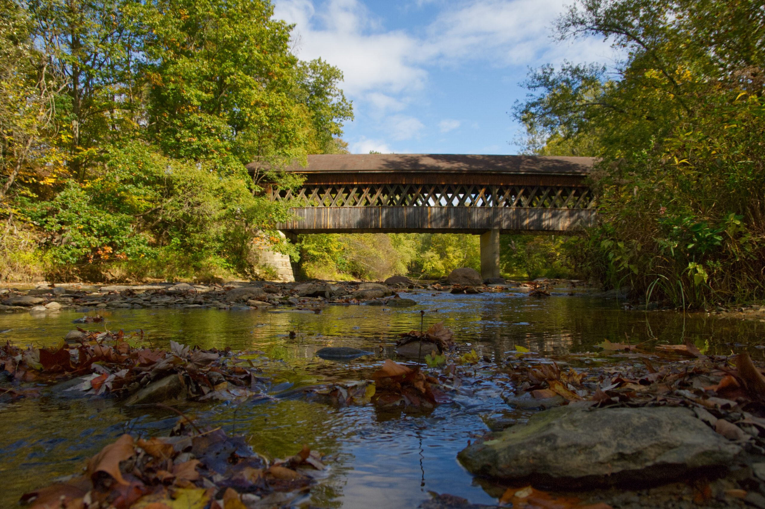 State road bridge in ashtabula viewed at the side from the river. There are fall leaves, a blue sky, and rocks in the river.