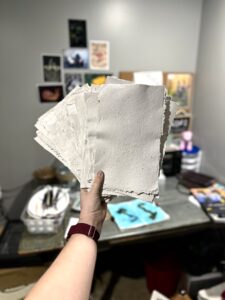 Holding up hand made paper in front of a table with painting supplies